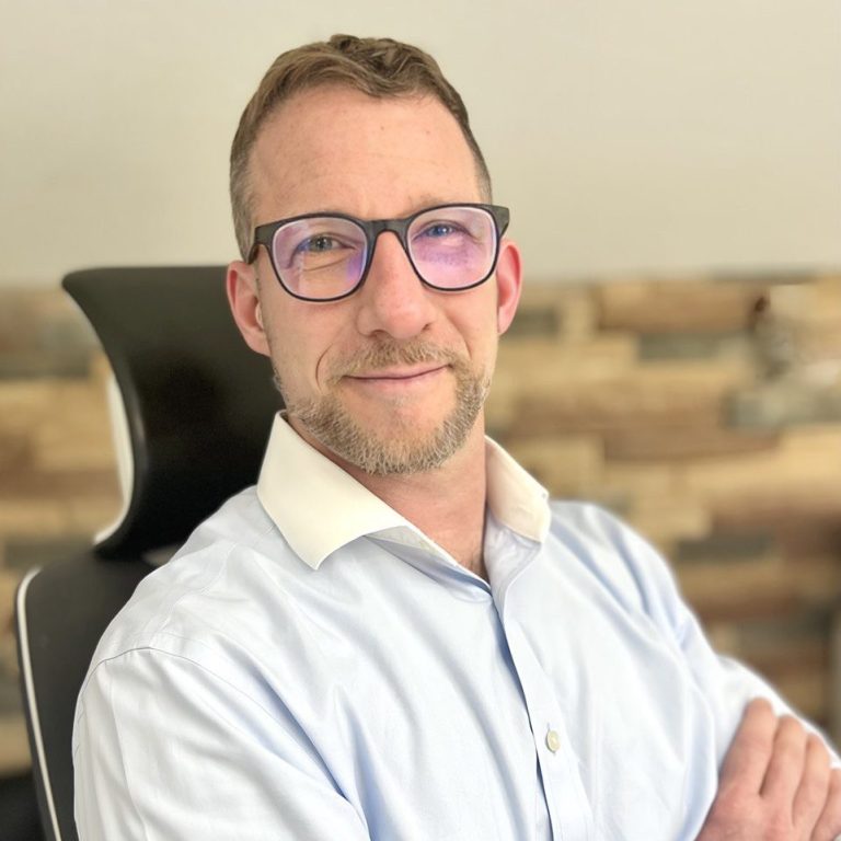 Markus Schierz - Steuerberater & Diplom-Betriebswirt (FH) Mann mit Brille, in blauem Hemd und lächelnd, vor einer Steinwand sitzend.